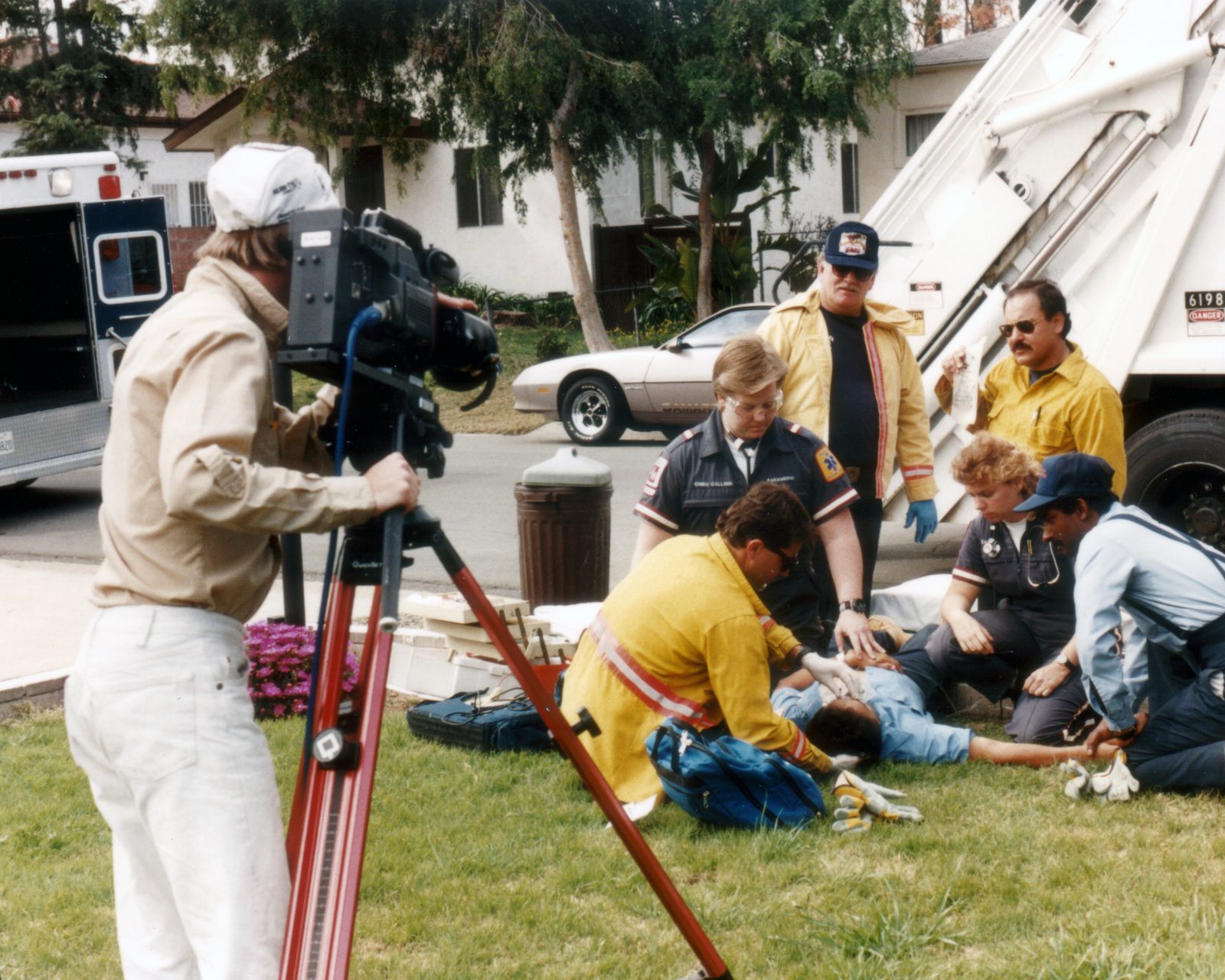 Videographer filming paramedics and firefighters tending to a sanitation worker in a dramatized emergency beside a garbage truck
