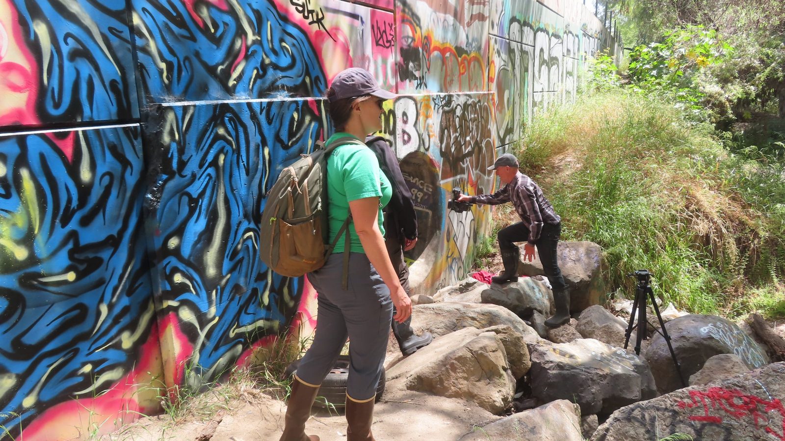Mark Schulze filming beside a graffiti-covered wall in the San Diego River bed with a San Diego River Park Foundation staff member